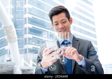 Giovane bell'uomo d'affari asiatico che usa lo smartphone all'aperto toccando lo schermo con interfaccia digitale virtuale che mostra le icone delle applicazioni Foto Stock