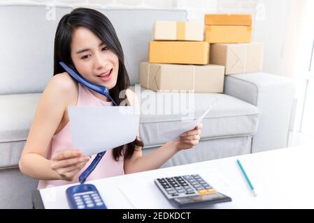 Giovane imprenditore asiatico di donna o venditore online freelance che lavora a. home conferma ordini da parte del cliente al telefono per la preparazione consegna Foto Stock