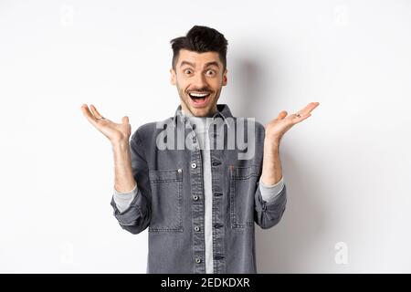 Un giovane eccitato che si acclama di buone notizie, si allarga le mani lateralmente e guarda felice, sorridente stupito, in piedi su sfondo bianco Foto Stock