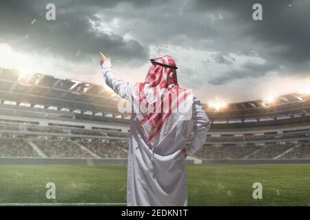 Uomo d'affari arabo vista posteriore in piedi sul suo stadio durante la partita sportiva. Stile di vita saudita moderno. L'uomo in abbigliamento tradizionale sembra sicuro, occupato. Etnia, finanza, reddito uncrease, sport, immobiliare Foto Stock