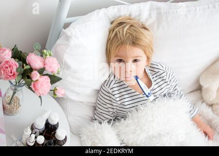 Malato piccolo biondo bambino, sdraiato a letto con febbre, abbracciando piccolo peloso giocattolo, medicina accanto a lui sul comodino Foto Stock