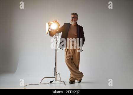 Foto a tutta lunghezza di un elegante uomo di mezza età in occhiali da vista, in piedi accanto al riflettore dello studio mentre si posa per la fotocamera su sfondo bianco. Foto di moda, concetto di stile Foto Stock