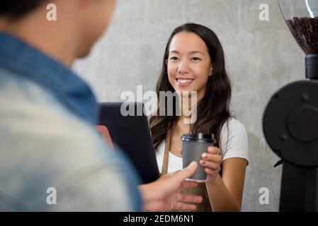 Il personale giovane e cordiale della donna asiatica offre una tazza di take-away caffè al cliente al banco del caffè Foto Stock