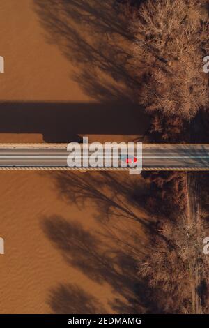 Vista aerea di una sola auto rossa che attraversa il ponte sul fiume, direttamente sopra il drone pov Foto Stock
