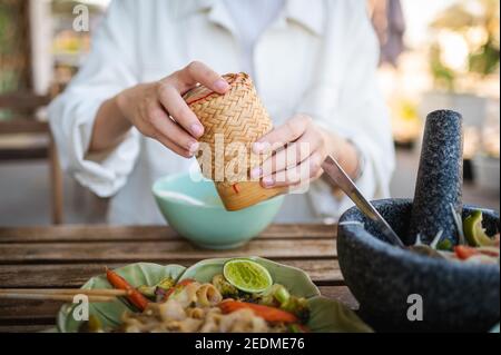 Donna che apre contenitore di bambù appiccicoso di riso mentre ha tre Pasto di portata in un guardaroba di ristorante tailandese Foto Stock