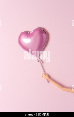 Mano di una bambola con palloncino a forma di cuore su sfondo rosa. Amore concetto minimo. Foto Stock