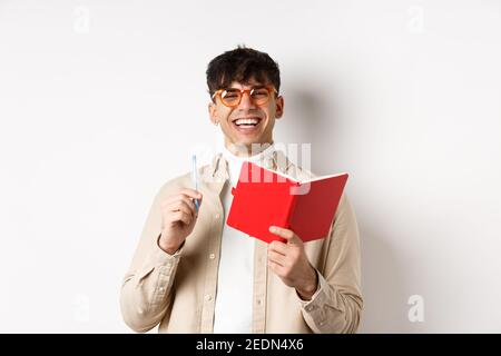 Giovane allegro in occhiali ridendo e prendendo appunti, scrivendo nel pianificatore, tenendo penna e diario, in piedi su sfondo bianco Foto Stock