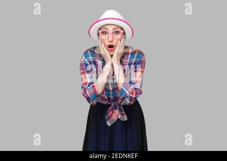 Ritratto di scioccato elegante donna matura in stile casual con cappello e occhiali in piedi, toccando il suo volto e guardando la telecamera con amazed faccia. ho Foto Stock