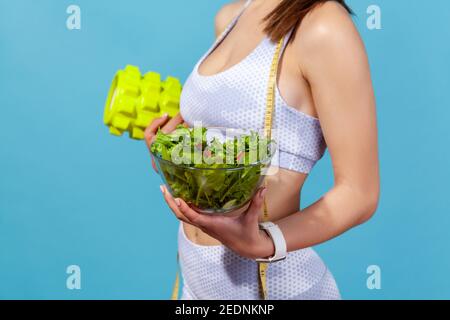 Primo piano vista laterale donna in abbigliamento sportivo bianco con metro a nastro che mostra la ciotola con insalata di verdure fresche e massaggiatore a rullo, programma di dimagrimento. Foto Stock