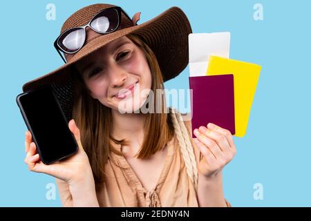 Ritratto di caucasica ragazza felice in estate cappello mostrando smartphone, documenti e la carta di vaccinazione alla fotocamera su uno sfondo blu. Concetto di viaggio Foto Stock