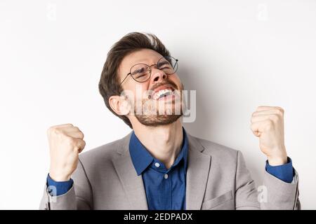 Uomo d'affari eccitato in occhiali e vestito gridando sì con piacere e volto sollevato, scuotendo pugni in su, trionfando, scommessa vincente, in piedi su bianco Foto Stock