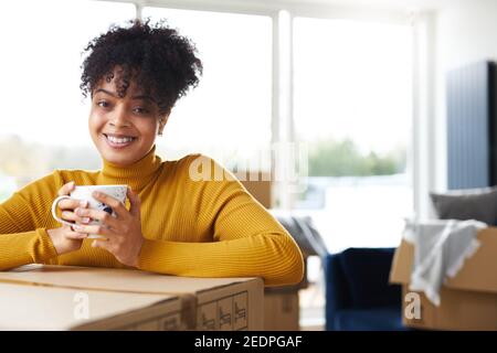 Ritratto di giovane donna fiduciosa in salotto di nuova casa riposandosi sulla scatola di rimozione e bevendo il caffè durante il giorno in movimento Foto Stock