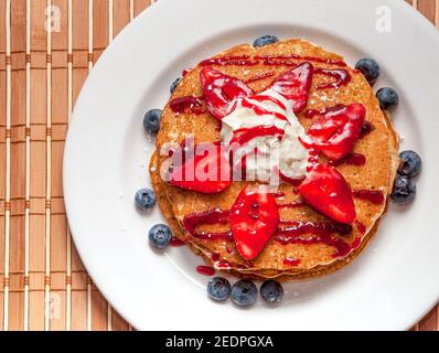 Frittelle con le bacche. Una pila corta di frittelle si siede su una piastra bianca, conditi con panna montata, fragole, mirtilli e sciroppo di fragole. Foto Stock