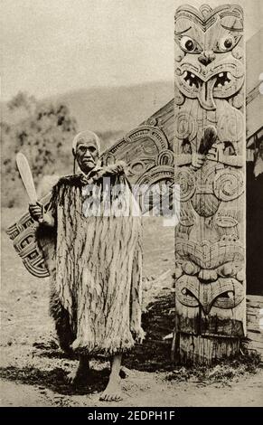 Una foto degli inizi del XX secolo di un anziano Maori che indossa A Korowai un mantello tradizionale fatto di fibre di lino He porta anche un patu pounamu o semplicemente un club fatto da pounamu greenstone si trova di fronte ad un wharenui o una sala riunioni scolpita Foto Stock