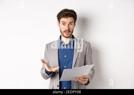 Uomo d'affari confuso che guarda il documento difettoso, lamentandosi sull'affare, indicando la carta frustrato, levandosi in piedi nel vestito su sfondo bianco Foto Stock