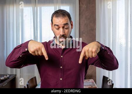 Bell'uomo bearned in una camicia viola che punta con i due forefingers delle sue mani giù in sorpresa. Concetto di sorpresa Foto Stock