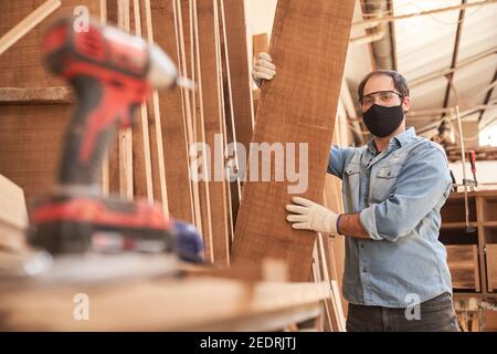 Artigiano come falegname con maschera a causa di Covid-19 nel negozio di legno del falegname Foto Stock