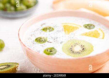 Yogurt con kiwi, uva spina, chia in ciotola di ceramica su fondo grigio di cemento e tessuto di lino arancione. Vista laterale, primo piano, messa a fuoco selettiva. Foto Stock