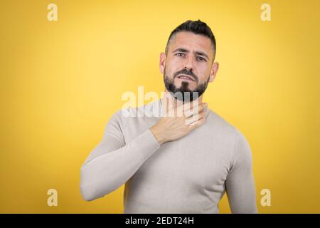 Bell'uomo con barba che indossa un maglione su sfondo giallo che tocca il collo doloroso, mal di gola per influenza, coaguli e infezioni Foto Stock