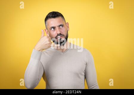 Bell'uomo con barba che indossa un maglione su sfondo giallo confuso fare il telefono con le mani e le dita come parlare il telefono Foto Stock