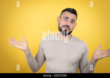 Bell'uomo con barba che indossa un pullover su sfondo giallo senza clueless e l'espressione confusa con braccia e mani sollevate Foto Stock