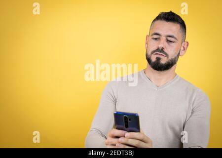 Bell'uomo con barba che indossa un pullover su uno sfondo giallo con il suo telefono Foto Stock