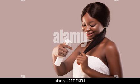 Sorridente Black Lady applicando spray idratante per capelli, facendo Split finisce trattamento di riparazione Foto Stock