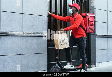 Pilota africano che consegna il pasto ai clienti con scooter elettrico mentre indossa la maschera facciale durante l'epidemia di virus corona Foto Stock