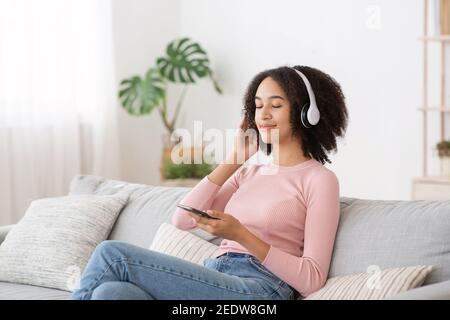 Donna divertente felice con le cuffie si siede in un comodo divano, ascoltando nuova musica pop Foto Stock