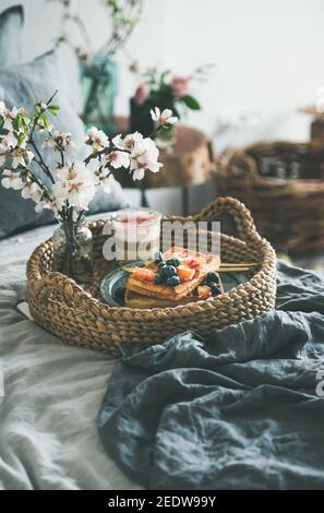 Prima colazione in camera. Waffle con arancio e mirtilli di sangue, latte di rosa in vetro e fiore bianco in vassoio cesto su biancheria da letto di lino grigio scuro, camera interna sullo sfondo, fuoco selettivo Foto Stock