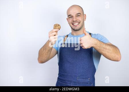 Giovane uomo calvo con grembiule uniforme su isolato sfondo bianco Giovane uomo calvo con grembiule uniforme su isolato sfondo bianco tenendo una choc Foto Stock