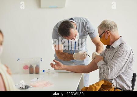 Vista laterale ritratto del medico maschile che vaccina l'uomo anziano indossando maschera in clinica medica, copia spazio Foto Stock