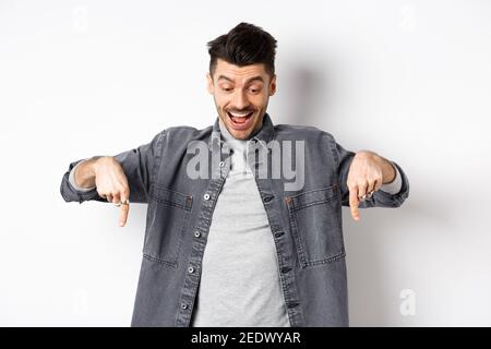 Un uomo europeo eccitato che guarda e punta le dita verso il basso, guardando felice alla pubblicità, in piedi in denim giacca su sfondo bianco Foto Stock