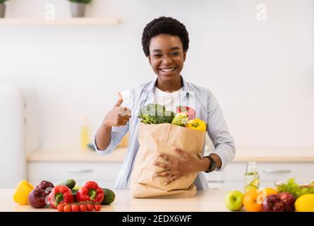 Black Woman gesturing Thumbs-Up Holding Grocery Shopping Bag in cucina Foto Stock