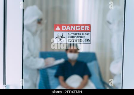 Closeup dell'etichetta con avviso di pericolo con focolaio di covid-19 messaggio sulla porta avere una giovane paziente femminile con i medici Vestita con kit DPI che prende appunti sui progressi compiuti Foto Stock