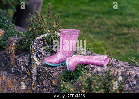 Un paio di stivali wellington in gomma glittery rosa per bambini abbandonati su un muro. Un montante e uno sul lato. Foto Stock