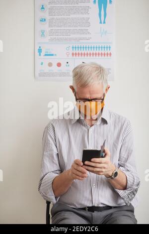 Ritratto verticale di un uomo anziano con capelli bianchi che indossa una maschera e. utilizzo dello smartphone durante l'attesa in linea presso la clinica medica Foto Stock
