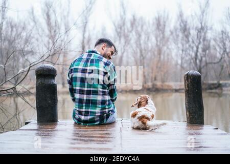 Sorridente uomo ispanico con flanella e cavalier re Charles spaniel su un molo in legno nel lago Foto Stock