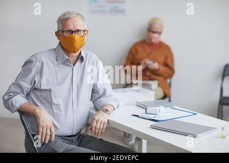 Ritratto di uomo anziano che indossa la maschera e guardando la macchina fotografica mentre si siede alla scrivania presso l'ufficio dei medici, copy space Foto Stock