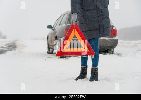 Donna mette in primo piano un cartello di avvertimento triangolare su una strada in una giornata invernale innevata. Concetto di guasto dell'auto Foto Stock