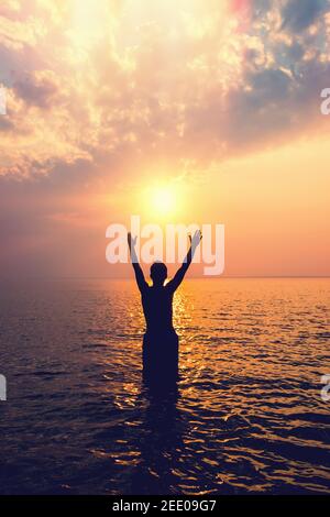 Toned Photo of Man Silhouette with Hands Up in the Acqua sullo sfondo del tramonto Foto Stock