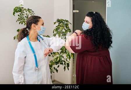Donna sovrappeso e il suo medico indossando maschere mediche si salutano l'un l'altro da gomito che urta presso l'ufficio del medico. Gomito a gomito, battaglia coronavirus epidemia Foto Stock
