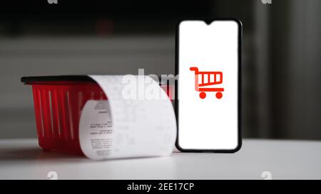 Icona del carrello sullo schermo dello smartphone accanto al cestino della spesa rosso e alla ricevuta della spesa. Online Store concetto di shopping e commerce addict shopping Foto Stock