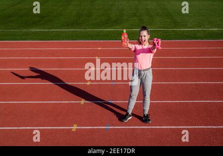 ragazza adolescente che tiene barbells e bottiglia d'acqua. allenatore di fitness prepararsi per l'allenamento. riscaldamento sulla palestra dello stadio. potenza e forza. allenamento dei bambini a scuola lezione di educazione fisica. manubri. Foto Stock