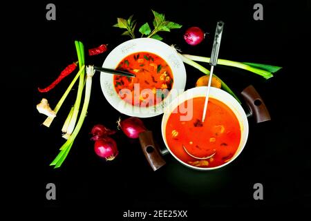 Zuppa piccante con carne di pollo, erbe e verdure su piatto bianco e in pentola, verdure ed erbe in disposizione intorno, su fondo nero. Foto Stock