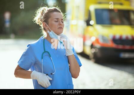 pandemia di coronavirus. donna paramedica moderna stanca in scrub con stetoscopio e maschera medica che respira all'esterno vicino all'ambulanza. Foto Stock