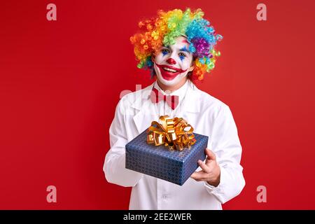 clown divertente con una scatola regalo isolato su sfondo rosso, giovane harlequin guardando la macchina fotografica Foto Stock