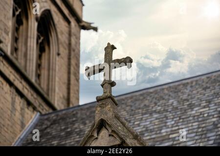 Sandstone old weathered stone religious cross on christian church building roof over ancient chapel door with tower and cloudy storm sky above sun Foto Stock