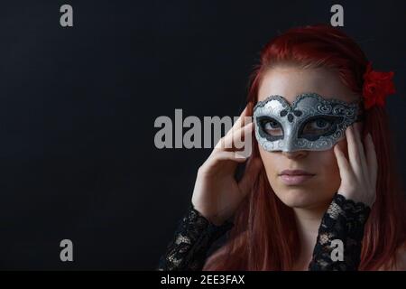 Basso ritratto chiave di giovane donna rossa tenendo in mano argento carnevale maschera sul viso. Orizzontalmente. Foto Stock