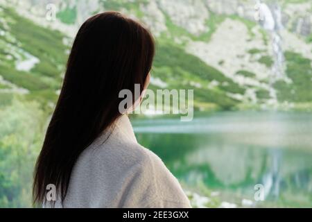 Giovane donna in un accappatoio bianco guardando nella finestra con vista mozzafiato sulle montagne e la cascata. CONCETTO DI SPA e benessere. Foto Stock